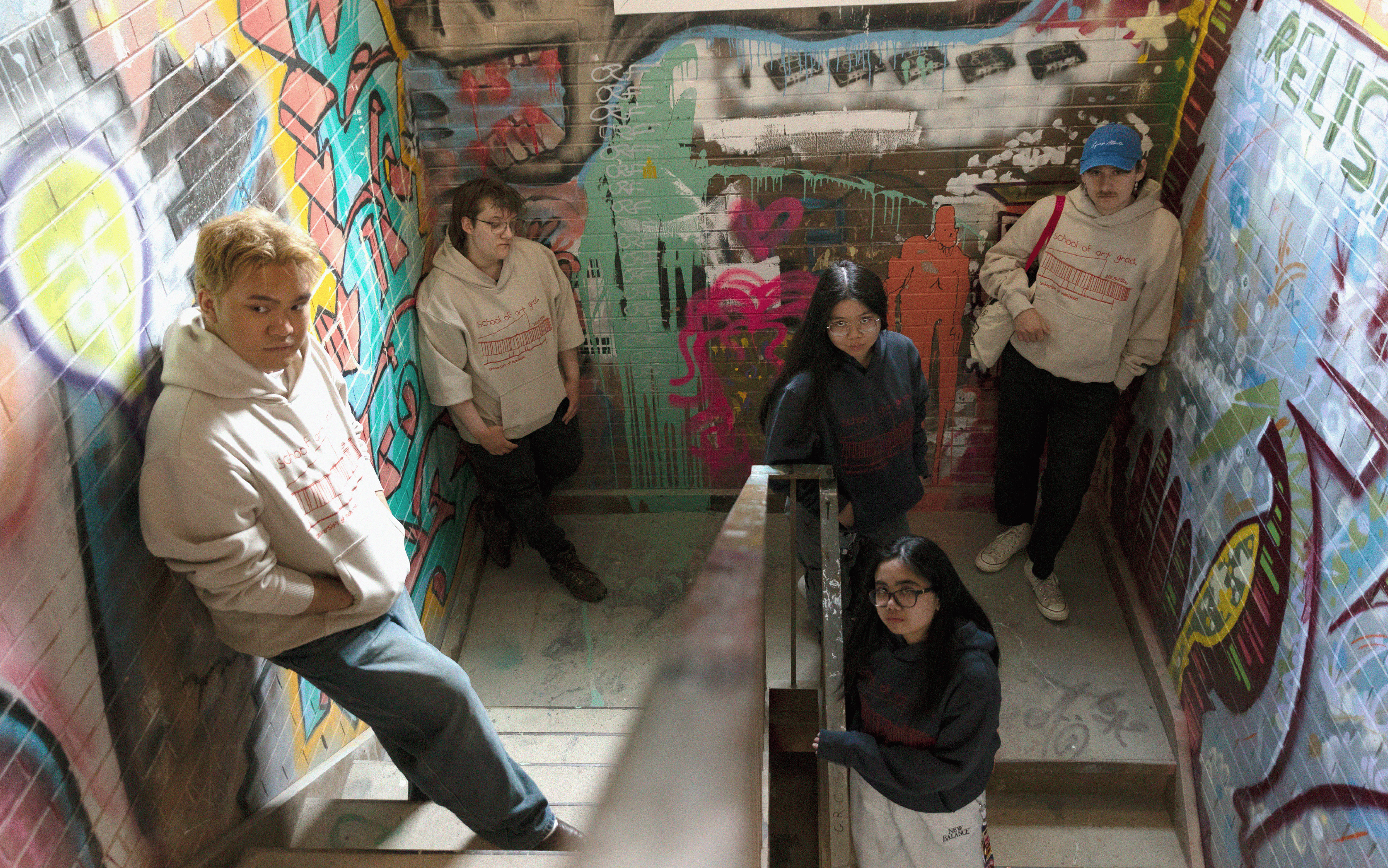 Students wearing graduation hoodies in a graffiti-covered stairwell.